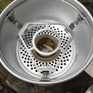Könnte beinhalten: Ein silberfarbener Campingkocher aus Metall mit einer kreisförmigen Basis, die zahlreiche Belüftungslöcher aufweist. Eine dreieckige Metallstützstruktur befindet sich in der Mitte und ist mit dem Text KARIBOU OUTDOORS versehen.