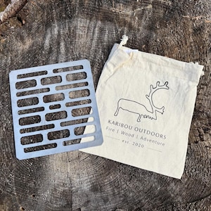 Può includere: Una griglia per barbecue in acciaio inossidabile con un design rettangolare e una serie di ritagli rettangolari, accanto a un sacchetto di tela con una grafica di renna e il testo "KARIBOU OUTDOORS Fire | Wood | Adventure est. 2020".