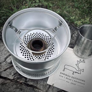 Może przedstawiać: Srebrna aluminiowa kuchenka kempingowa z okrągłą perforowaną podstawą i centralnym palnikiem, w zestawie z miarką ze stali nierdzewnej oraz białym płóciennym workiem z czarnym logo renifera i napisem KARIBOU OUTDOORS Fire | Wood | Adventure est. 2020.