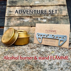 Könnte beinhalten: Ein goldfarbenes Alkoholbrenner- und Ständerset. Das Set enthält einen kleinen, runden Brenner mit Deckel und einen silberfarbenen Ständer. Die Texte "ADVENTURE SET" und "FLÄMME" sind sichtbar.