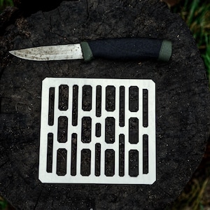 Könnte beinhalten: Ein weißes Camping-Gitter aus Metall mit einem Messer mit schwarzem Griff und grünem Akzent auf einem dunkelbraunen Baumstumpf.