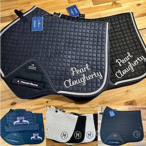 May include: Three different saddle pads with embroidered names. The first saddle pad is navy blue with white piping and the name "Pearl Clougherty" embroidered in white. The second saddle pad is black with white piping and the name "Pearl Clougherty" embroidered in white. The third saddle pad is black with white piping and a gear design embroidered in white.