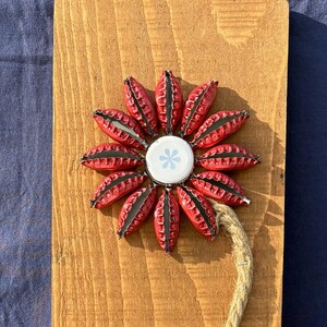 May include: A handcrafted flower made from red bottle caps, with a white and blue center, mounted on a wooden plank. The flower has a jute rope stem. The wooden plank has a natural wood grain.