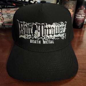 Può includere: Un cappellino da baseball nero con testo ricamato in bianco sul pannello frontale. Il ricamo mostra le parole Bolt Thrower in un carattere stilizzato con le parole DEATH METAL stampate in un carattere più piccolo e semplice sotto il logo.