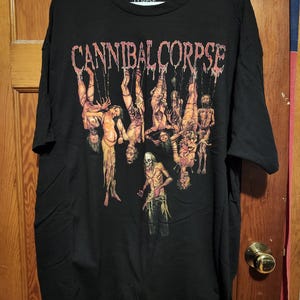 Puede incluir: Una camiseta negra de manga corta con el texto Cannibal Corpse en una fuente roja estilizada en la parte superior. Debajo del texto hay una ilustración gráfica de varias figuras colgadas de cuerdas, con un esqueleto de pie en la parte inferior.