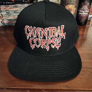 May include: A black baseball cap featuring the text CANNIBAL CORPSE embroidered in red with a white outline. The lettering has a dripping, liquid-like effect. The cap has a structured crown and a flat brim, maintaining a solid black colour throughout.