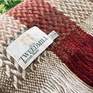 Può includere: Primo piano di una coperta tessuta con un'etichetta che recita "TWEEDMILL British Made". La coperta presenta un mix di colori rosso, marrone e crema con un motivo strutturato e bordi sfrangiati. L'etichetta include anche il testo "Est. 1971".