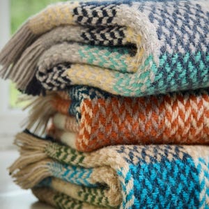 May include: A stack of three folded woven blankets featuring a herringbone pattern. The top blanket is blue, gray, and yellow. The middle blanket is orange and cream. The bottom blanket is blue, green, and cream. Each blanket has fringed edges.