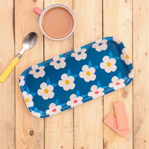Puede incluir: Una bandeja rectangular azul con un estampado floral blanco, una taza con asa rosa con té, una cuchara con mango amarillo y obleas rosas sobre una superficie de madera. La bandeja mide unos 30 cm de largo.