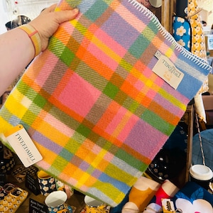 Puede incluir: Una manta a cuadros colorida en tonos rosa, naranja, amarillo, verde y azul. La manta tiene un borde cosido blanco y una etiqueta que dice "TWEEDMILL". La manta se sostiene frente a una exhibición de otros artículos.