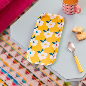 May include: A yellow metal tray with a white floral pattern. The tray is rectangular and has a slightly rounded edge. The flowers are stylized and have blue and red accents.