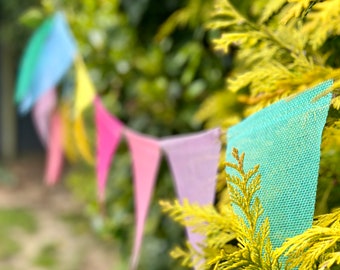 May include: A string of colorful bunting flags hanging from a tree branch. The flags are made of a textured fabric and are in shades of blue, yellow, pink, and purple.