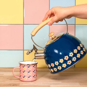 May include: A blue enamel tea kettle with a wooden handle and a floral design is pouring into a pink floral mug. The kettle has a silver spout and a wooden knob on the lid. The mug is on a wooden surface.