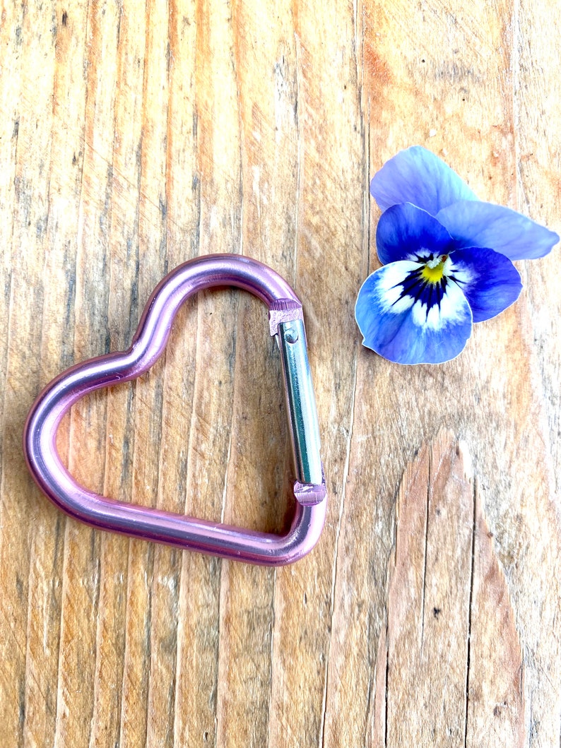 May include: A pink heart-shaped carabiner clip on a wooden surface with a purple and white pansy flower.
