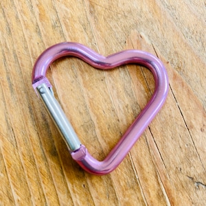 May include: A pink heart-shaped metal carabiner with a silver spring-loaded gate.