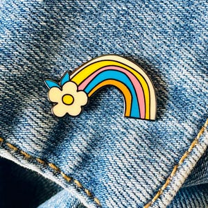 May include: A rainbow-shaped enamel pin in yellow, blue, orange, and pink, with a white flower and blue leaves. The pin is set against a blue denim background. The pin is a decorative accessory.