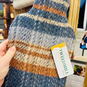 May include: A rectangular hot water bottle cover with a woven pattern in blue, brown, and cream. The cover has a tag that reads "TWEEDMILL". The product is designed to hold a hot water bottle.