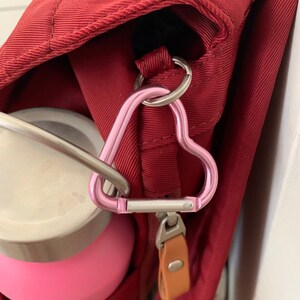 May include: A red bag with a pink heart-shaped carabiner attached to a zipper pull. A pink water bottle is attached to the carabiner.