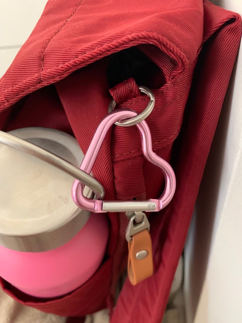 May include: A red bag with a pink heart-shaped carabiner attached to a zipper pull. A pink water bottle is attached to the carabiner.