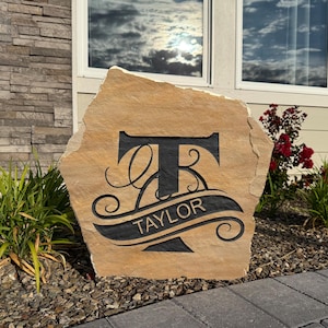 May include: A tan, irregularly shaped natural stone garden marker featuring a large, black, engraved capital letter T. A black banner curves across the center of the letter, containing the word TAYLOR in a clean, sans-serif font.
