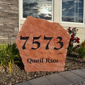 May include: A large, irregular, reddish-orange natural stone address marker featuring the engraved black numbers 7573 and the text Quail Run. The stone has a rough, textured surface and is positioned upright in a garden setting.