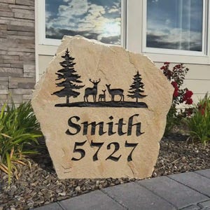 May include: A tan, irregular-shaped stone garden sign featuring a black engraved design of two pine trees and three deer. Below the nature scene, the text Smith and the numbers 5727 are deeply carved into the stone surface in a dark, bold font.
