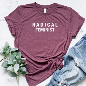 Puede incluir: Una camiseta de color granate con el texto "RADICAL FEMINIST" impreso en blanco en la parte delantera. La camiseta est&aacute; colocada sobre una superficie de m&aacute;rmol blanco con un par de vaqueros azules y una rama de follaje verde.