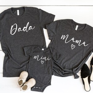 May include: Three gray t-shirts with white text. The largest shirt says "Dada", the medium shirt says "Mama" with a heart, and the smallest shirt says "mini" with a heart.