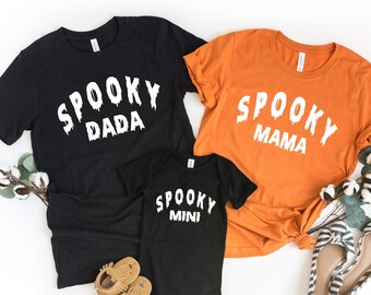 May include: Three black and orange t-shirts with the text "Spooky Dada", "Spooky Mama", and "Spooky Mini" in white dripping letters. The shirts are laid out on a white background with cotton branches and a pair of black and white striped shoes.
