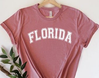 May include: A dusty rose t-shirt with the word "FLORIDA" printed in white on the front. The shirt is on a hanger and has rolled up sleeves.