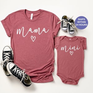 May include: A set of two shirts in a dusty rose color. The larger shirt has the word "mama" written in white with a small heart. The smaller shirt has the word "mini" written in white with a small heart.
