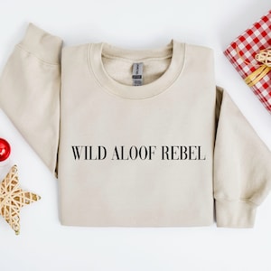 Peut inclure: Un sweat-shirt beige avec le texte "WILD ALOOF REBEL" imprimé sur le devant. Le sweat-shirt est plié et posé à plat sur une surface blanche.