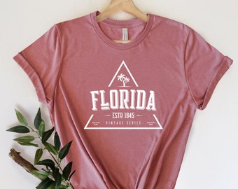 May include: A dusty rose t-shirt with a white graphic design of a triangle with a palm tree inside. The text "Florida - Est. 1845 - Vintage Series" is inside the triangle.