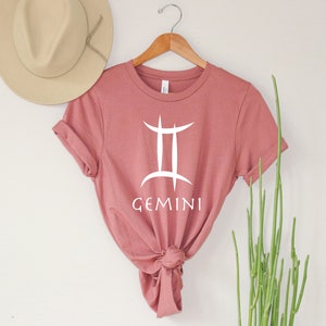 May include: A dusty rose t-shirt with a white Gemini zodiac symbol and the word "GEMINI" printed on the front.