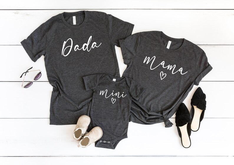 May include: Three gray t-shirts with white text. The largest shirt says "Dada", the medium shirt says "Mama" with a heart, and the smallest shirt says "mini" with a heart.