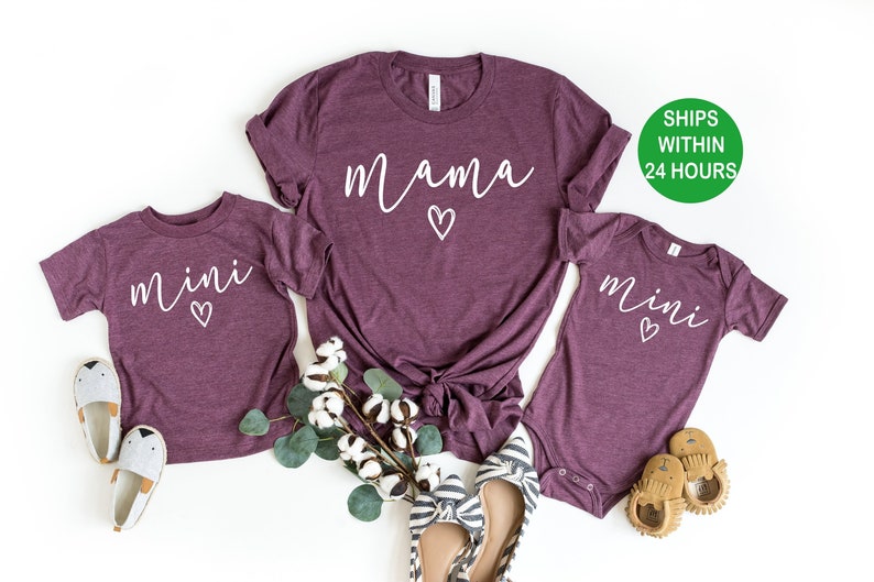May include: A set of three maroon t-shirts with white text. The largest shirt says "mama" with a small heart. The two smaller shirts say "mini" with a small heart.