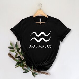 Puede incluir: Camiseta negra con un signo del zod&iacute;aco Acuario blanco y la palabra "Acuario" impresa en la parte delantera.