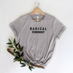 Puede incluir: Una camiseta gris con el texto "RADICAL FEMINIST" impreso en negro en la parte delantera.