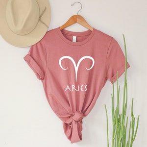 Puede incluir: Una camiseta rosa polvorienta con un s&iacute;mbolo del zod&iacute;aco Aries blanco y la palabra "ARIES" impresa en la parte delantera.