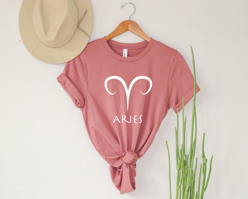 Puede incluir: Una camiseta rosa polvorienta con un s&iacute;mbolo del zod&iacute;aco Aries blanco y la palabra "ARIES" impresa en la parte delantera.