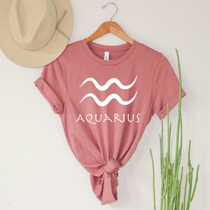 Puede incluir: Una camiseta rosa polvorienta con un gr&aacute;fico blanco del s&iacute;mbolo astrol&oacute;gico de Acuario y la palabra "Aquarius" debajo.