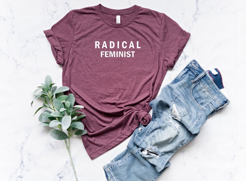 Puede incluir: Una camiseta de color granate con el texto "RADICAL FEMINIST" impreso en blanco en la parte delantera. La camiseta est&aacute; colocada sobre una superficie de m&aacute;rmol blanco con un par de vaqueros azules y una rama de follaje verde.