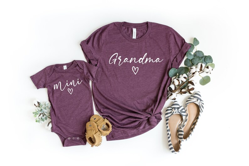 May include: A set of two maroon t-shirts with white text. The larger shirt says "Grandma" with a small heart. The smaller shirt says "mini" with a small heart.