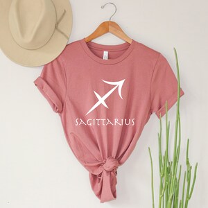 May include: A dusty rose t-shirt with a white Sagittarius zodiac symbol and the word "Sagittarius" printed on the front.