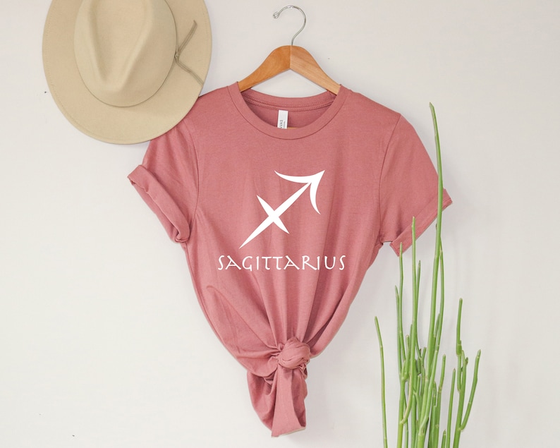 May include: A dusty rose t-shirt with a white Sagittarius zodiac symbol and the word "Sagittarius" printed on the front.