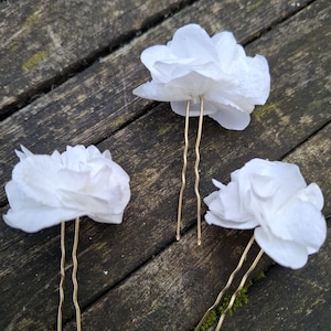 Pode incluir: Três grampos de cabelo de flores de tecido branco com pinos dourados. As flores são sobrepostas e têm uma textura delicada e enrugada.