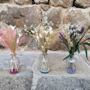 Op de afbeelding: Drie glazen vazen met gedroogde bloemen. De vaas links heeft roze en witte gedroogde bloemen. De middelste vaas heeft witte en beige gedroogde bloemen. De vaas rechts heeft paarse en witte gedroogde bloemen.