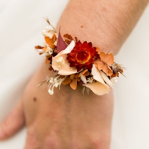 Peut inclure: Un bracelet floral composé de fleurs séchées dans des tons rouge profond, orange brûlé et crème. L'arrangement comprend une immortelle rouge centrale, des pétales d'hortensia orange et de délicats brins de gypsophile blancs sur un bracelet.