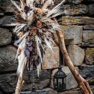 Pode incluir: Duas lanternas de metal preto penduradas em um galho de madeira rústico contra uma parede de pedra. O galho é decorado com flores secas e capim-dos-pampas.