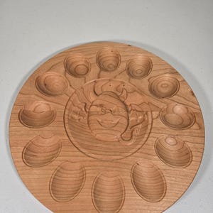 May include: A round, wooden serving tray with a carved design. The tray features a central circular carving of a smiling face, surrounded by 12 carved depressions for holding items. The wood has a natural, light brown color.